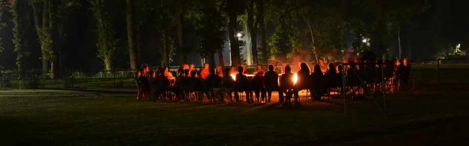 Kinderen zitten samen rond het kampvuur onder hoge bomen.