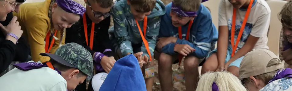 Kinderen zitten lachend in een kring rond een kampactiviteit.