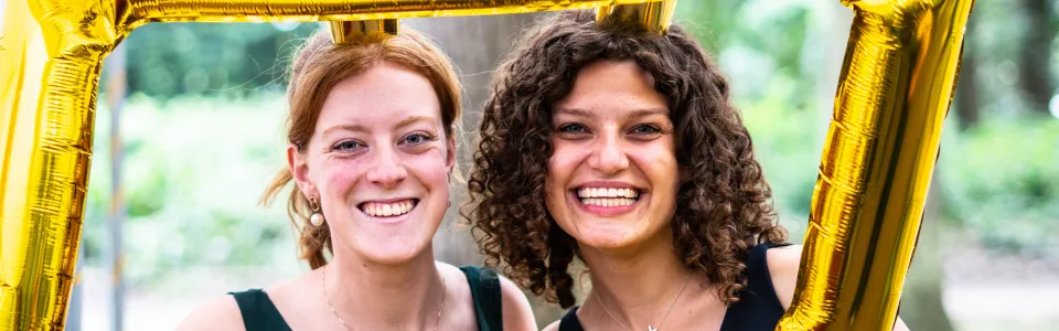 Twee lachende kampleidsters poseren achter een gouden ballonlijst