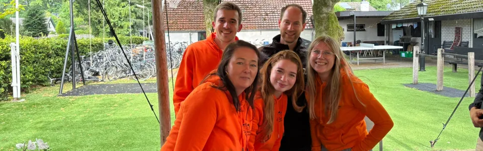 Vijf kampbegeleiders poseren lachend in oranje truien onder de tent.